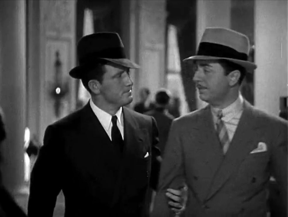 Two men in suits and hats walk closely, conversing in an elegant indoor setting. The scene is in black and white, suggesting a classic era.  Spencer Tracy and William Powell in the Hollywood film screwball 1930s comedy classic movie “Libeled Lady”