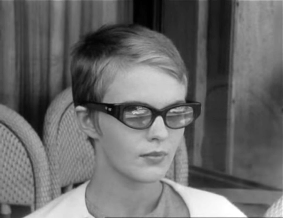 A person with short hair and sunglasses sits calmly on a chair. The background features wicker furniture and curtains, evoking a serene mood. Michel's car is reflected in Jean Seberg's sunglasses in the classic French New Wave film 1960 romantic crime drama thriller movie by Jean-Luc Godard "Breathless"