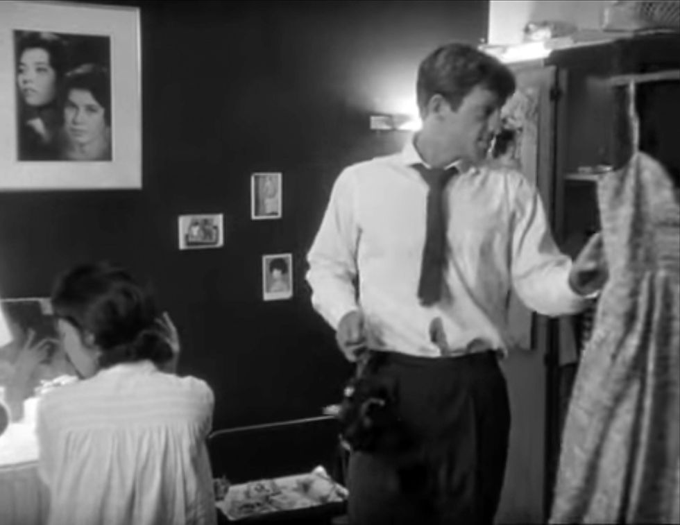 Man in tie holds dress, woman applies makeup at vanity. Black-and-white room with photos on wall, calm mood. Lilian Dreyfus and Jean-Paul Belmondo in the classic French New Wave film 1960 romantic crime drama thriller movie by Jean-Luc Godard "Breathless"