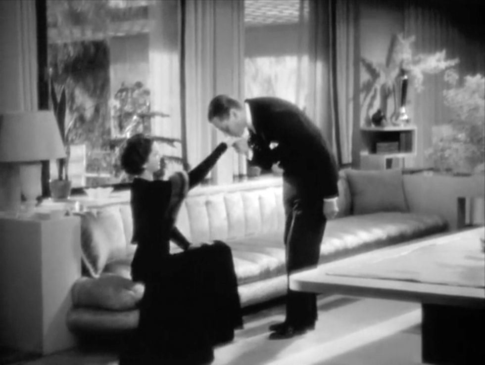 A man in a suit bows and kisses a woman's hand in a classic living room with a long sofa and curtains. The scene is elegant and formal. Herbert Marshall kisses Kay Francis hand in her art deco apartment in the classic Ernst Lubitsch romantic comedy movie 1932 Hollywood film "Trouble in Paradise"