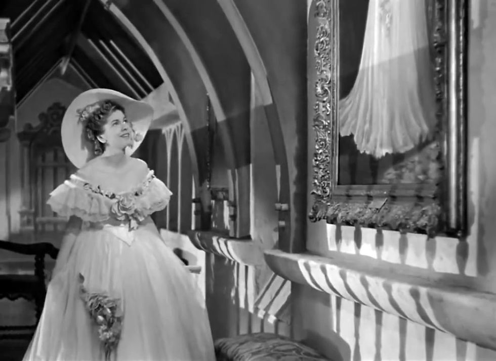 Woman in a frilly dress and wide hat looks joyfully at a framed gown. Gothic-style arched hallway with intricate shadows. Black and white. Joan Fontaine duplicates a dress from a painting in the classic Alfred Hitchcock suspense movie film "Rebecca"