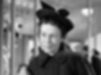 Woman in a dark coat and hat stands indoors, looking contemplative. Blurred background includes lights and shadows. Black and white image. Thelma Ritter as a mom in the classic movie Hollywood holiday Christmas Santa Claus film "Miracle on 34th Street"