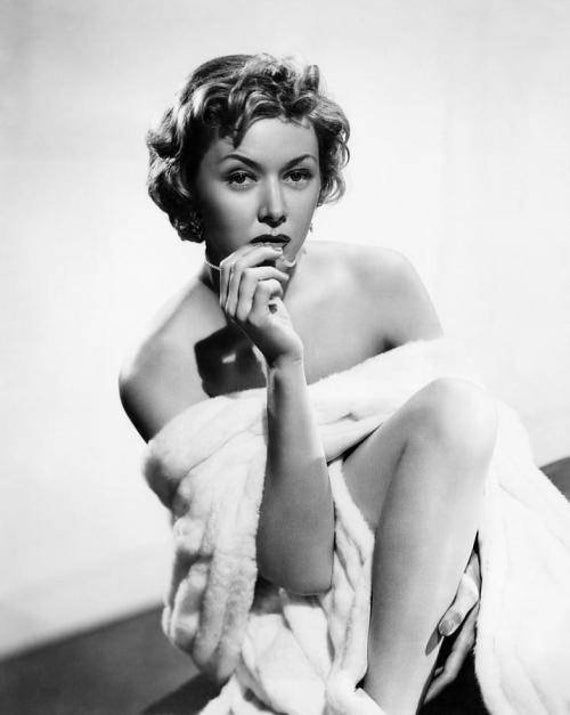 Woman in a fur wrap poses thoughtfully on a stool against a plain white background. Black and white photograph exudes a vintage, reflective mood. Glamour portrait of Hollywood movie star film actress smoldering young Gloria Grahame