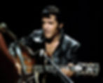 A person in a black leather jacket plays a guitar, singing into a microphone on stage. The background is dark, highlighting the performance. Color portrait photo of young King of Rock and Roll movie star singer icon legend Elvis Presley from his 1968 Comeback holiday speical