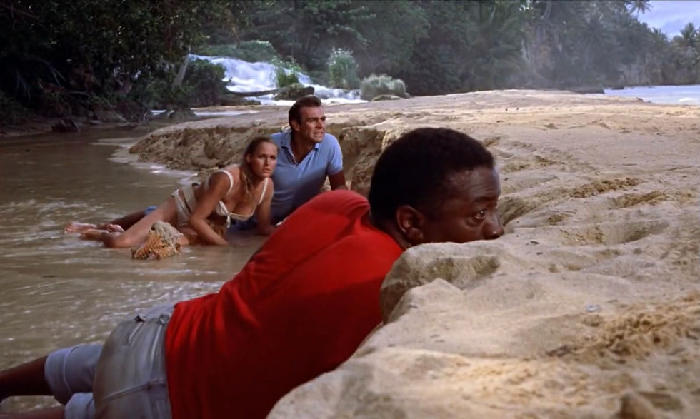 Three people crouch in shallow river water by rocky waterfalls, looking tense and alert in a tropical jungle setting. Ursula Andress, Sean Connery and John Kitzmiller hide in the sand on Dr. No's island Crab Key in the first classic James Bond movie espionage spy thriller 007 film 1962 "Dr. No"