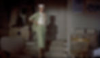 Woman in green suit and white hat stands pensively in a vintage living room. Beige armchair, wooden chair, and stacks of papers visible. Grace Kelly shows off a women's suit designed by Edith Head in the Alfred Hitchcock classic Hollywood film suspense voyeuristic movie "Rear Window" Peeping Tom