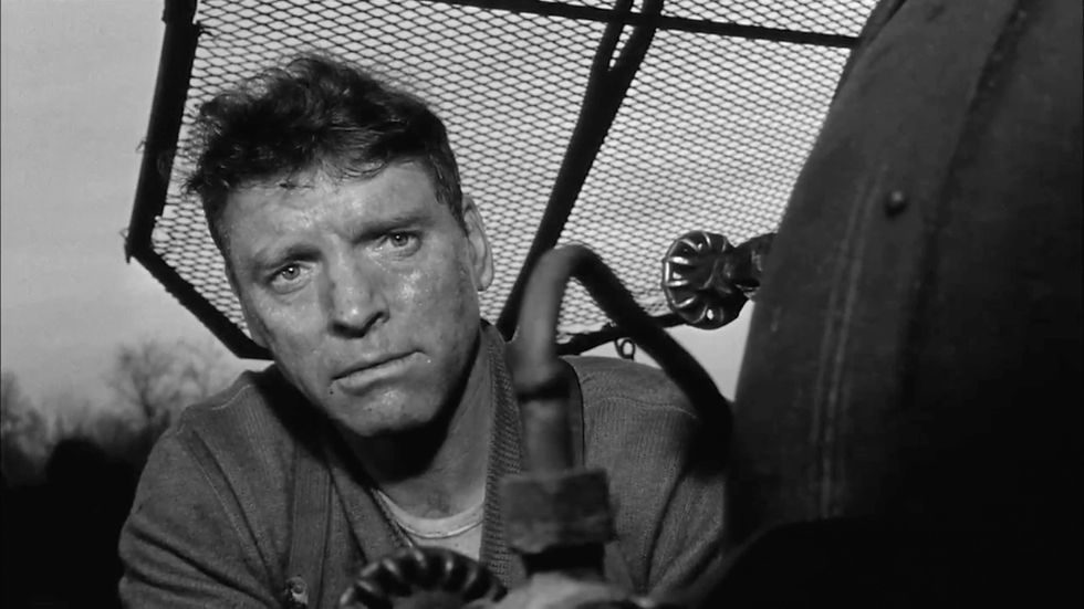 A man looks intensely at the camera, with a machine propeller in the foreground. The setting is outdoors with a mesh fence in the background. Burt Lancaster drives the tain in a scene from the 1964 action adventure movie World War II WWII film about the Nazis and stolen art, "The Train"