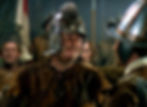Medieval armored men face off in a dim hall, one smiling in a dented helmet, others blurred behind them. Alan Hale as an Irish rebel in the classic Hollywood costume drama 1939 movie about Queen Elizabeth I and the Earl of Essex in "The Private Lives of Elizabeth and Essex"
