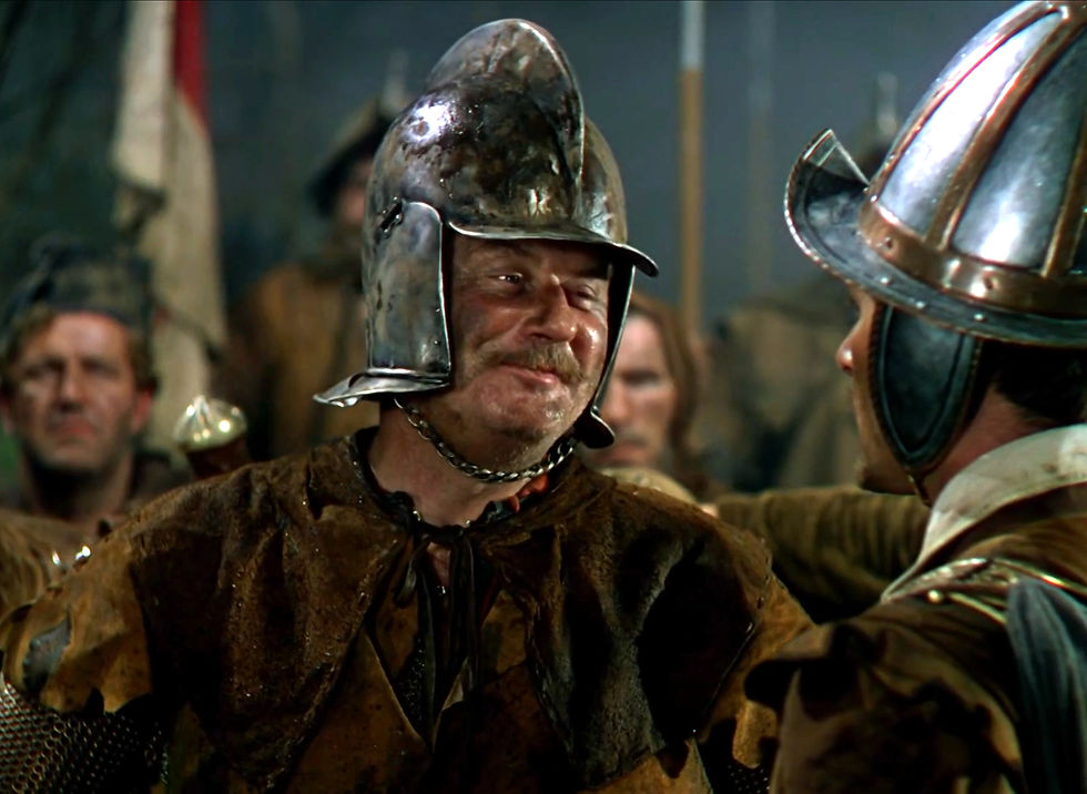 Medieval armored men face off in a dim hall, one smiling in a dented helmet, others blurred behind them. Alan Hale as an Irish rebel in the classic Hollywood costume drama 1939 movie about Queen Elizabeth I and the Earl of Essex in "The Private Lives of Elizabeth and Essex"