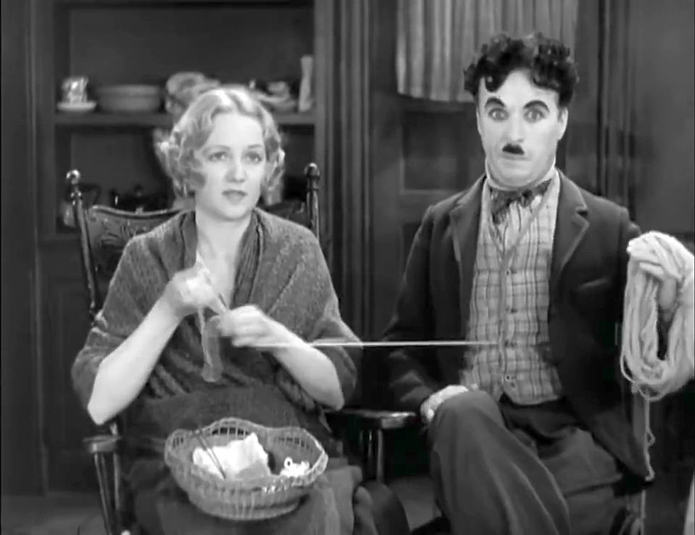 Woman knitting and man holding yarn in a vintage room. She appears focused, he looks humorous. Shelves with dishes in the background. Black and white. Virginia Cherrill and Charlie Charles Chaplin in the classic Hollywood silent movie City Lights