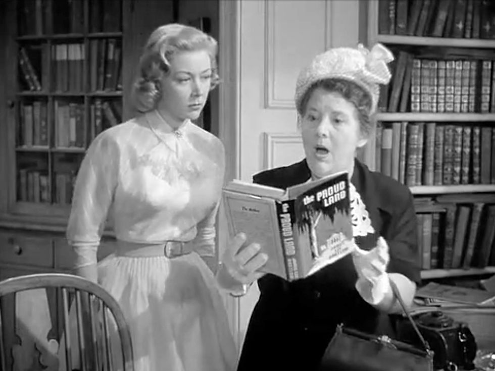 Two women in a library, one reading "The Proud Land" aloud with surprise. Bookshelves in the background, creating a tense mood. Madge Blake reads her book inscription as Gloria Grahame looks on in the classic 1952 melodrama film about Hollywood moviemaking movies "The Bad and the Beautiful"