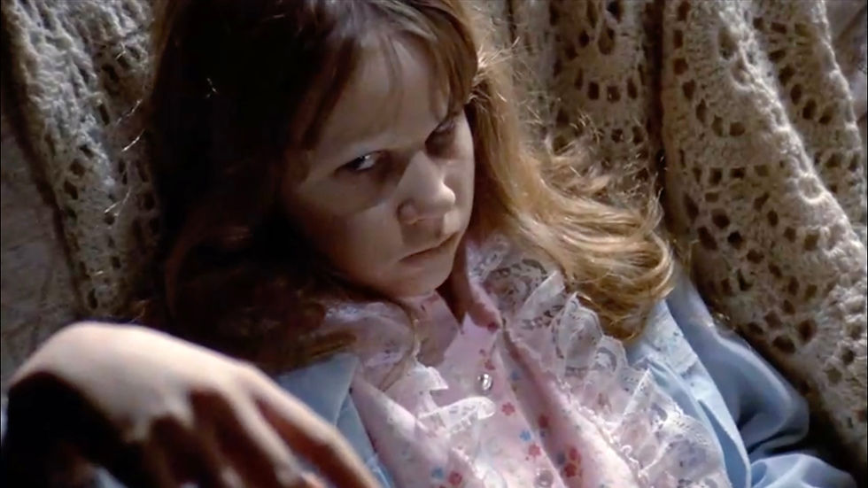 Young girl in a pink lace bib and blue top stares sternly at the camera, framed by a crocheted blanket background. Linda Blair as "Regan" possessed by the devil demon Pazuzu in the scariest movie of all time, classic horror film about the devil and possession, 1973 "The Exorcist".