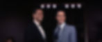 Two men in suits smile on stage under bright lights. One wears black and white, the other in a light blue suit. Dark background adds contrast. Cesare Danova and Elvis Presley in the classic 1960s Elvis Presley rocking MGM movie musical "Viva Las Vegas".