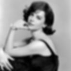 Woman in a black dress poses elegantly against a gray background. She wears a floral bracelet, pearl earrings, and looks serene. Hollywood movie star film actress portrait photo of young Natalie Wood