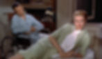 A man in a wheelchair in blue pajamas and a woman in a green suit on a couch; they appear relaxed in a cozy room with a vintage phone. James Jimmy Stewart looks at Grace Kelly who lays on the bed in the Alfred Hitchcock classic Hollywood film suspense movie "Rear Window"