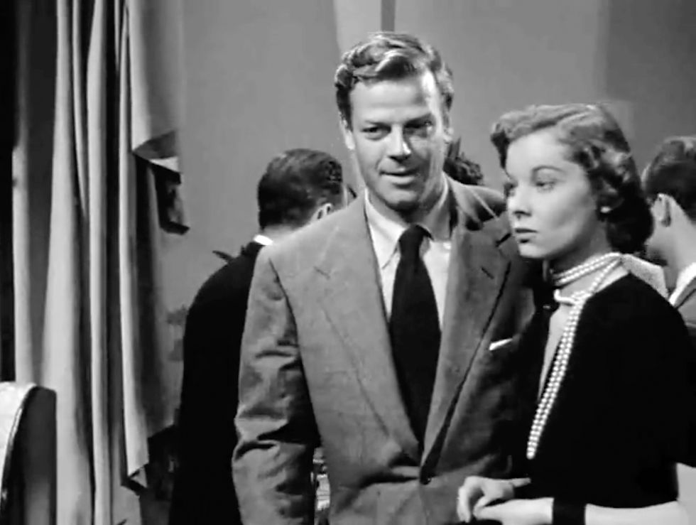 A man in a suit and a woman in a pearl necklace stand close in a dimly lit room. The mood is serious, with curtains in the background. James Arness has a small part in the classic Hollywood movie romance Nicholas Ray film noir "In a Lonely Place"