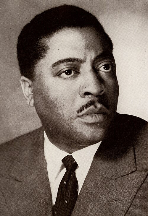 Black-and-white portrait of a serious man in a suit and tie, looking off to the side against a plain studio background. Portrait of Black American film actor European movie star young John Kitzmiller