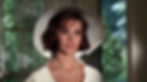 Woman in a white hat and dress, wearing a pearl necklace, standing indoors near a window with green foliage outside. She appears calm. Natalie Wood stars as Deanie Loomis in the classic 1960s Hollywood film romance movie about sex and repression, "Splendor in the Grass"