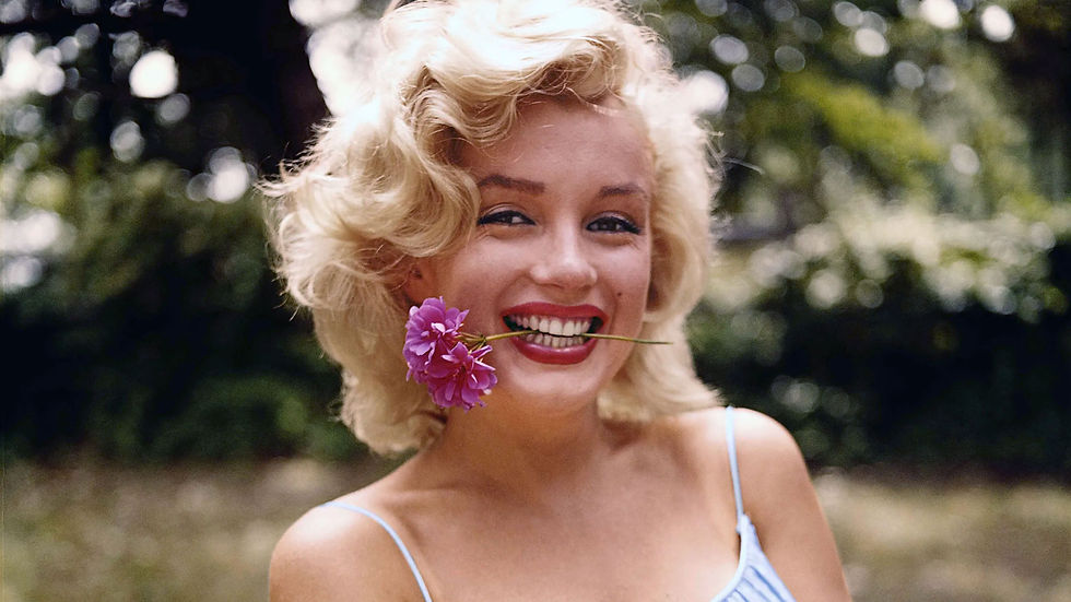 Smiling blonde woman outdoors holds purple flowers in her mouth, with blurred green trees behind her. Portrait of blonde Hollywood movie star sex symbol goddess icon legend Norma Jeane Baker actress Marilyn Monroe color photo younger with flower in her mouth