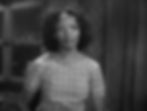 Woman with curly hair in a plaid dress stands indoors, looking forward with a neutral expression. Dimly lit background. No text visible. Black Hollywood classic character actress Theresa Harris in the Hollywood Pre-Code film sexy racy classic movie "Baby Face"