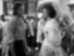 Man in worn shirt and woman in elegant dress with a large hat face each other, surrounded by onlookers, in a vintage setting. Black and white. Olivia de Havilland buys Errol Flynn at a slave aution to save his life in the classic 1930s swashbuckling pirate film classic Hollywood adventure movie "Captain Blood"