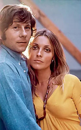 A man in a blue shirt and a woman in a yellow dress with a necklace pose closely. The background is neutral, creating a calm mood. Color portrait of young Polish international, Hollywood film writer movie director Roman Polanski with Hollywood movie star actress and wife Sharon Tate
