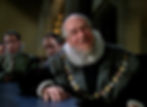 Older bearded man in ornate ruff and fur robe leans at a table in a candlelit hall, with stern men seated behind him. Henry Stephenson with Henry Daniell behind him in the classic Hollywood costume drama 1939 movie about Queen Elizabeth I and the Earl of Essex in "The Private Lives of Elizabeth and Essex"