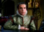 Serious man in ornate Renaissance dress sits at a table in a dim candlelit room, staring ahead. Henry Daniell in costume by Orry-Kelly in the classic Hollywood costume drama 1939 movie about Queen Elizabeth I and the Earl of Essex in "The Private Lives of Elizabeth and Essex"