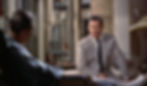 Two men converse in a dimly lit room. One in a gray suit sits on a sofa, looking concerned, while the other is in a chair facing away. Wendell talks with James Jimmy Stewart in the Alfred Hitchcock classic Hollywood film suspense voyeuristic movie "Rear Window" Peeping Tom