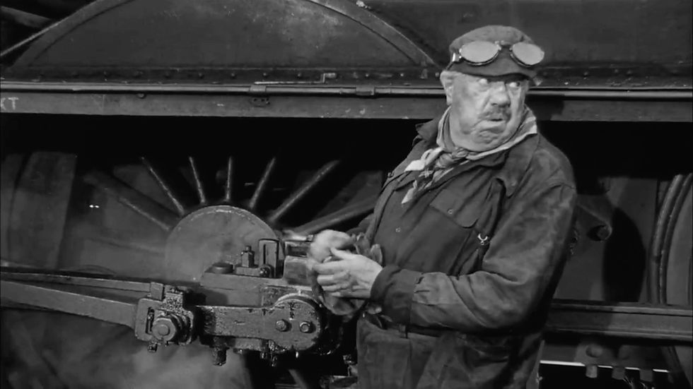 Elderly mechanic in overalls and goggles maintains a steam locomotive. He wipes his hands, standing near large wheels. Black-and-white setting. Michel Simon with the oil line in a scene from the 1964 action adventure movie World War II WWII film about the Nazis and stolen art, "The Train"