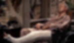 Man in a beige shirt asleep in a wheelchair, leg in a white cast. Dial phone and books on nearby table. Window view of trees. James Jimmy Stewart with a broken leg stars in the Alfred Hitchcock classic Hollywood film suspense movie "Rear Window"