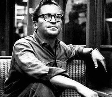 Man with glasses sits in a relaxed pose on a patterned bench, wearing a button-up shirt. Background is blurred. Mood is contemplative. Photo of Hollywood movie film, theater, Television director young Sidney Lumet on the subway