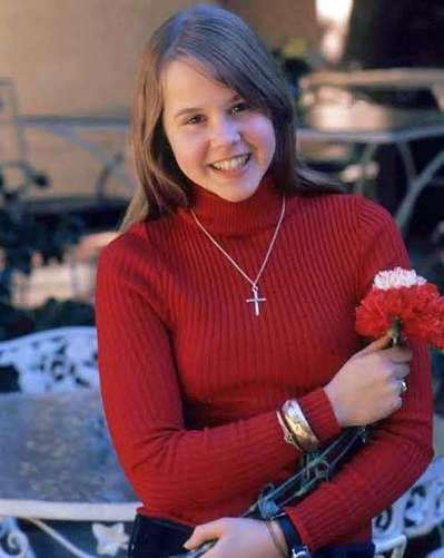 Smiling woman in a red sweater holding red flowers outdoors, wearing a cross necklace and bracelets. color photo of child star film horror icon Hollywood movie star actress TV young Linda Blair