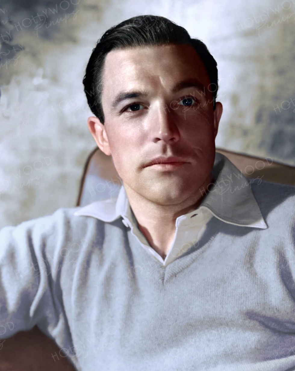 A man in a grey sweater and white shirt sits in a chair with a neutral expression. The background is a textured wall with faint text. Color portrait photo of young Hollywood movie star dancer choreographer Gene Kelly in light blue sweater