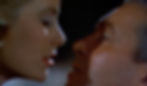 A close-up of a woman and man facing each other intimately, about to kiss. The background is dark, highlighting the faces. The mood is romantic. Grace Kelly kisses James Jimmy Stewart tenderly in the Alfred Hitchcock classic Hollywood film suspense voyeuristic movie "Rear Window" Peeping Tom