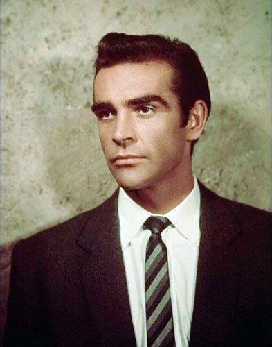 Portrait of a man in a dark suit and striped tie, looking off-camera against a mottled green background. Color portrait photo of Scottish movie star Hollywood film actor young James Bond Sean Connery 007