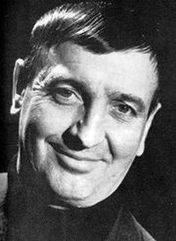 Smiling man in black-and-white photo, wearing a dark shirt. The background is dark, highlighting his cheerful expression. Portrait photo of French character film actor Hollywood movie star Albert Rémy