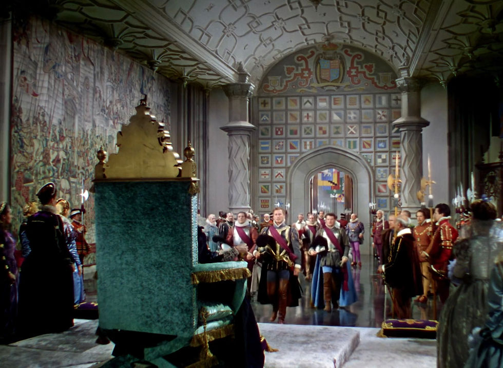 Courtiers in colorful medieval dress approach a throne in a grand tiled hall with arched doorway and royal crests. Bette Davis greets Errol Flynn in her throne at the palace in the classic Hollywood costume drama 1939 movie about Queen Elizabeth I and the Earl of Essex in "The Private Lives of Elizabeth and Essex"