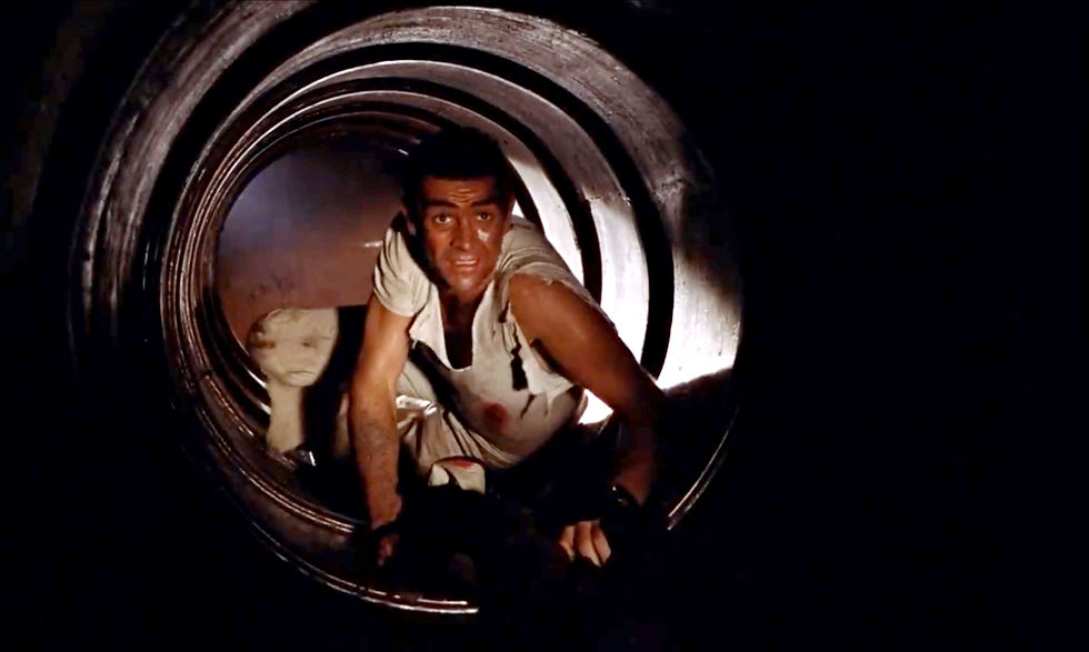 Man in a torn white shirt crawls through a dark metal pipe, lit from behind, with a tense, determined look. Sean Connery in the air vents in the first classic James Bond movie espionage spy thriller 007 film 1962 "Dr. No"
