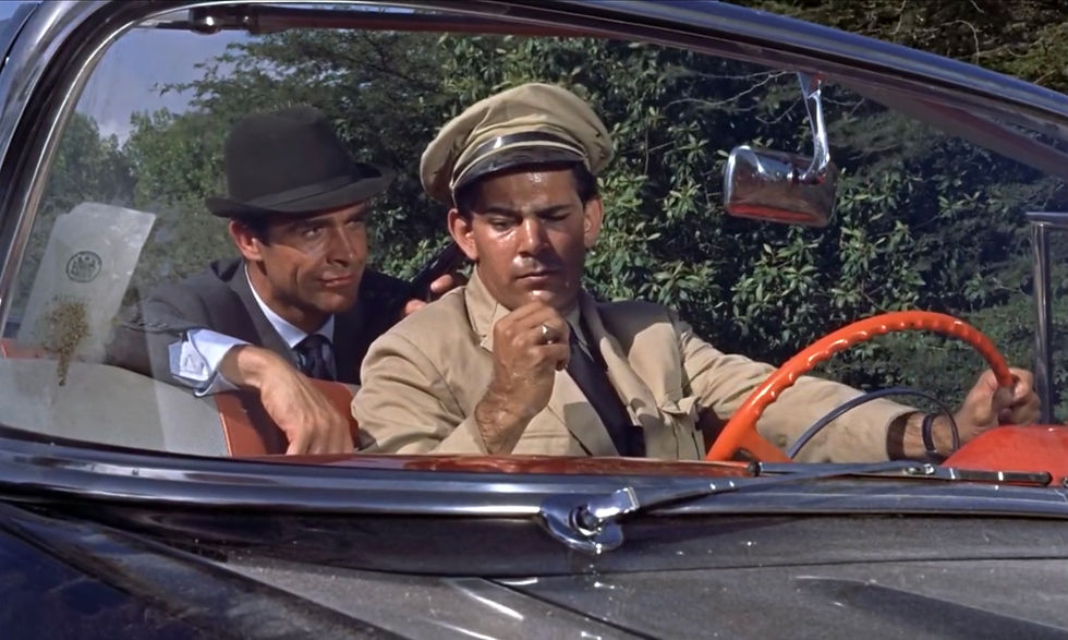 Two men in a vintage car, one driving tensely in a beige uniform cap while the other leans in close with a black hat; trees outside. Airport chauffeur Reginald Carter with Sean Connery in the first classic James Bond movie espionage spy thriller 007 film 1962 "Dr. No"