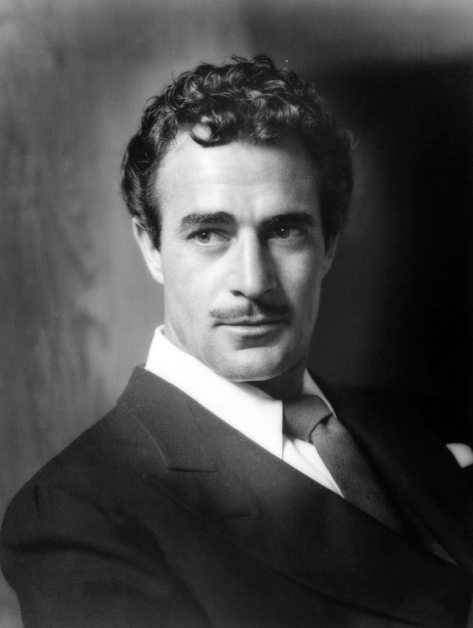 Black and white portrait of a man in a suit with a mustache, looking off-camera. Neutral expression, dark background enhances focus on face. George Hurrell portrait of Hollywood movie star Latin Lover handsome mustached film actor Mexican Gilbert Roland