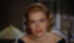 A woman with pearl earrings and necklace, red lipstick, wears a black dress, gazing warmly. Soft lighting, blurred background. Grace Kelly's entrance moving in for a kiss in the Alfred Hitchcock classic Hollywood film suspense voyeuristic movie "Rear Window" Peeping Tom