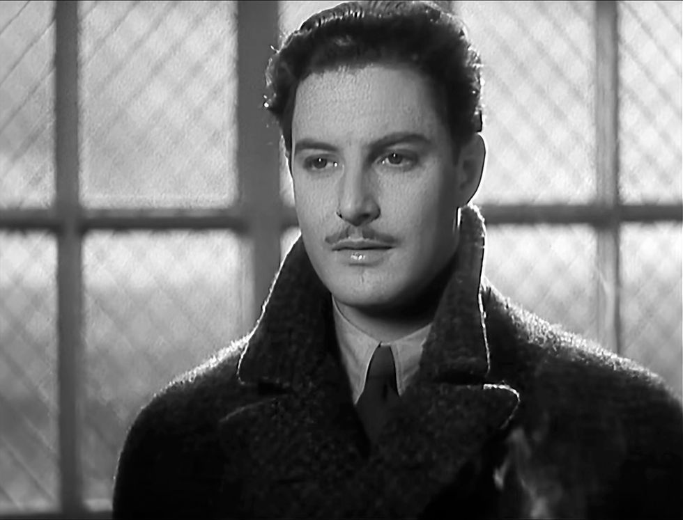 Black-and-white close-up of a serious man in a dark coat and tie, standing before a bright window with frosted panes. Robert Donat stars in the Alfred Hitchcock British film masterpiece spy thriller suspense movie "The 39 Steps"