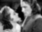 Two people in vintage attire gaze at each other intently in a black and white photo, conveying a dramatic and romantic mood. Olivia de Havilland and Errol Flynn star in the classic 1930s swashbuckling pirate film classic Hollywood adventure movie "Captain Blood"