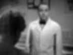 Man in a white coat looks surprised in a shop, with glass containers in the background. Blurred figure and sign with "Milk" partially visible. Black and white. Grady Sutton is a waiter in a southern diner in the classic Hollywood romantic comedy film rom-com movie 1940s "The More the Merrier".