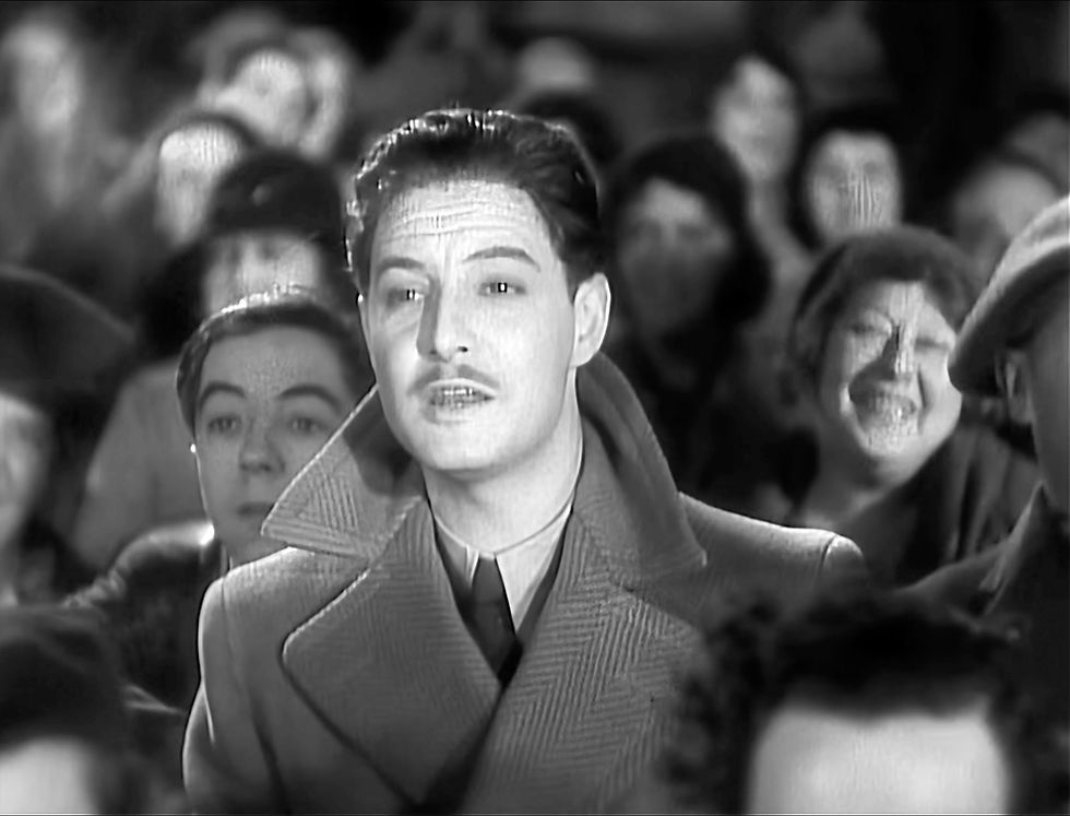 Black-and-white close-up of a man in a coat standing in a crowd, looking ahead with a serious, attentive expression. Robert Donat stars in the Alfred Hitchcock British film masterpiece spy thriller suspense movie "The 39 Steps"