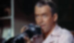 Man holding a camera with a large lens, focused expression. Wearing a light-colored shirt. Blurred background with muted tones. James Jimmy Stewart uses his camera's telephoto lens to watch his neighbors in the Alfred Hitchcock classic Hollywood film suspense voyeuristic movie "Rear Window" Peeping Tom