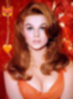 Woman with red hair in an orange dress against a vibrant red background. Heart-eye cat emoji and orange heart emoji on the left. Color portrait of young singing dancing Hollywood movie star Vegas entertainer TV film actress sex symbol kitten legend icon Ann-Margret