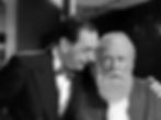 A man in a suit talks to a bearded man in a Santa outfit, inside a room with lockers. The bearded man looks surprised and concerned. Phillip Tonge and Edmund Gwenn in the classic movie Hollywood holiday Christmas Santa Claus film "Miracle on 34th Street"