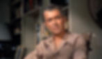 Man in a beige shirt sitting in a room with shelves of books. Neutral expression, warm lighting, cozy indoor setting. No visible text. James Jimmy Stewart stars in the Alfred Hitchcock classic Hollywood film suspense voyeuristic movie "Rear Window" Peeping Tom